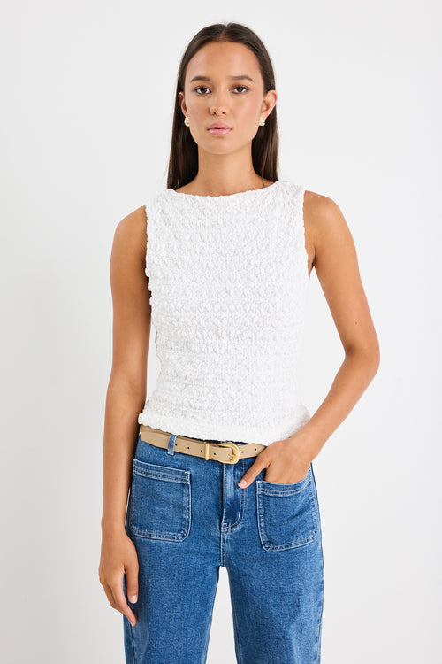 Woman wearing a white sleeveless textured top with a fitted silhouette and high neckline, paired with high-waisted denim jeans.