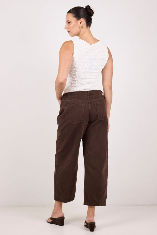 A woman shown from the back wearing a white textured sleeveless top and dark brown cropped trousers, standing against a plain light gray studio background.