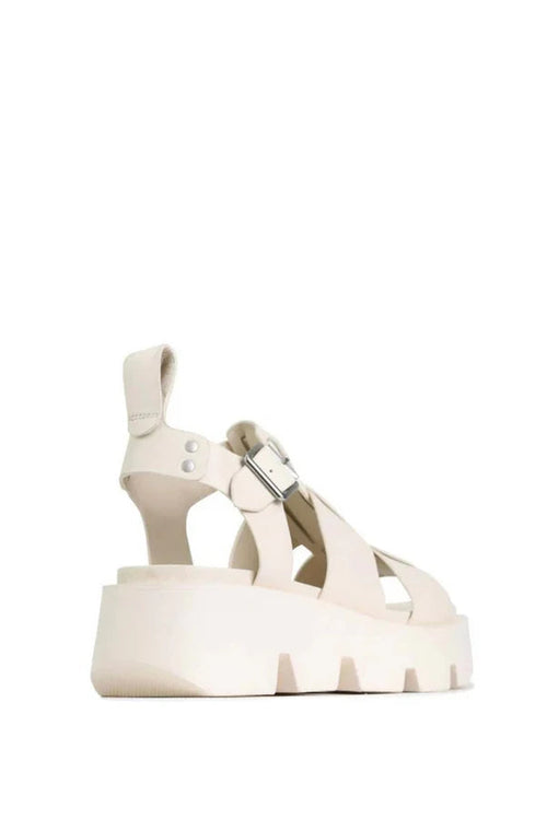 A single platform sandal in a light cream or off-white color, featuring a chunky sole, cutout straps, and a buckle detail on the ankle strap, shown against a plain white background.
