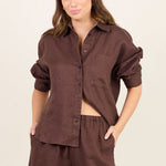 A woman modeling a matching dark brown lounge set consisting of a button-down shirt and shorts, paired with beige flip-flops, against a plain off-white studio background.