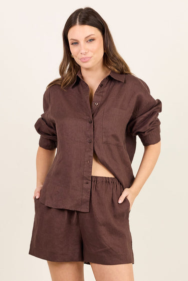 A woman modeling a matching dark brown lounge set consisting of a button-down shirt and shorts, paired with beige flip-flops, against a plain off-white studio background.