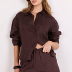 A woman modeling a dark brown two-piece linen set consisting of a button-down shirt and matching shorts, standing against a plain white background.