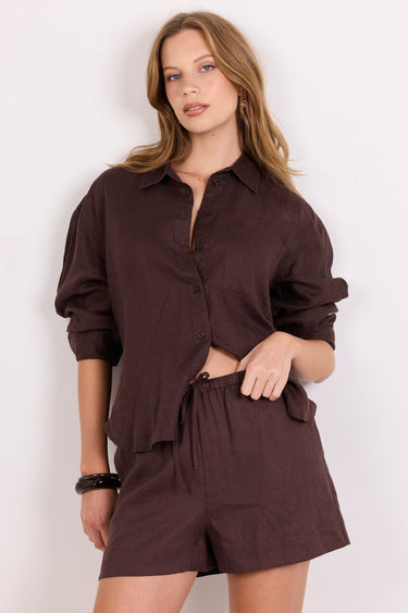 A woman modeling a dark brown two-piece linen set consisting of a button-down shirt and matching shorts, standing against a plain white background.