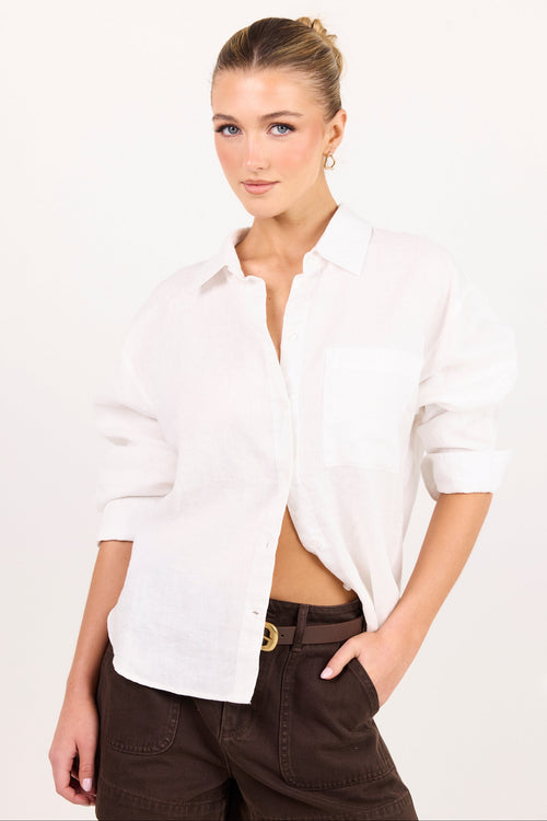 A woman modeling a white long-sleeve linen button-up shirt with rolled-up sleeves and dark brown pants, set against a plain white studio background.