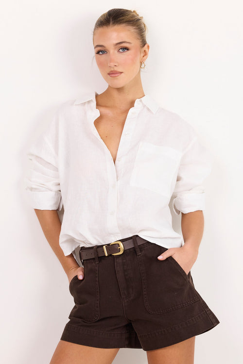 A woman modeling a white long-sleeve button-down shirt and dark brown shorts with a black belt, standing against a plain white background.