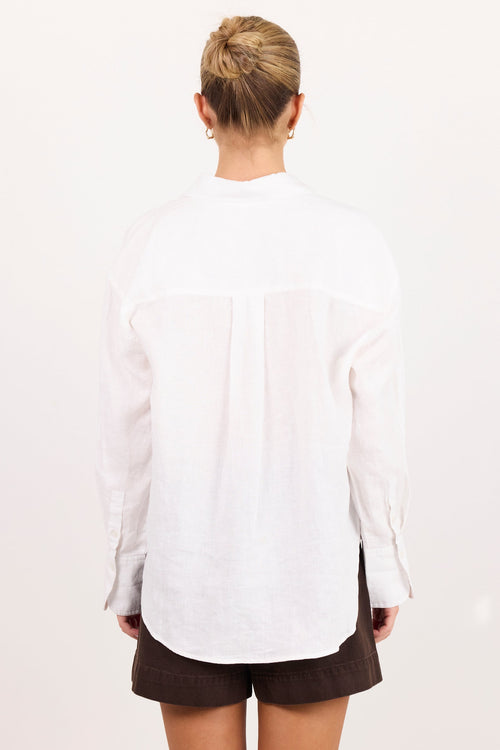 A woman viewed from behind wearing a white long-sleeve linen shirt and dark brown shorts against a plain white background.