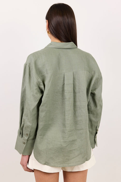 A woman viewed from the back wearing an oversized sage green linen button-up shirt and white shorts against a plain off-white background.