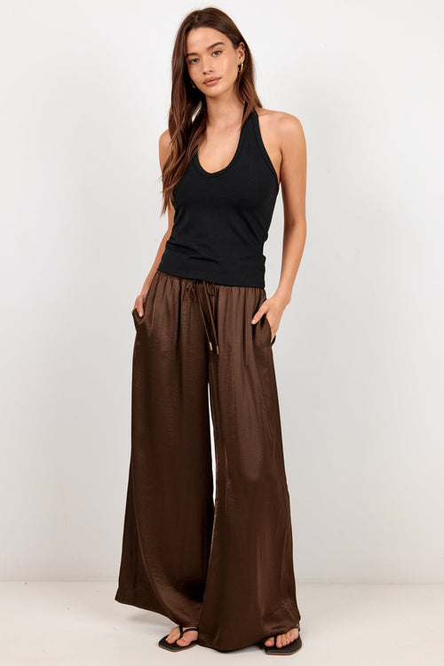 A woman modeling a black sleeveless tank top and wide-leg brown pants against a plain white studio background.