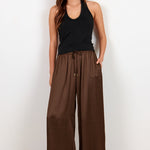A woman modeling wide-leg brown pants with a drawstring waist, paired with a black tank top and black sandals, standing against a plain white studio background.