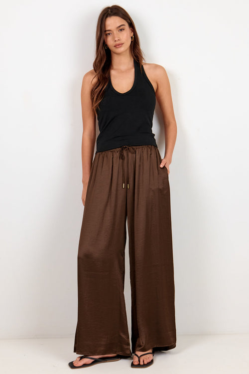 A woman modeling wide-leg brown pants with a drawstring waist, paired with a black tank top and black sandals, standing against a plain white studio background.