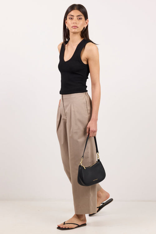 A woman modeling a black ribbed tank top, beige wide-leg cropped trousers, black sandals, and holding a black crescent-shaped shoulder bag with gold hardware, set against a plain light gray studio background.