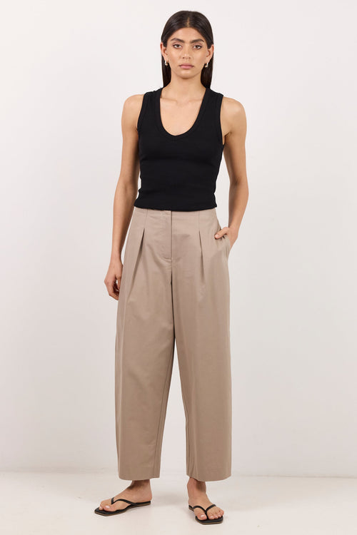 A model stands against a plain white studio background wearing a black sleeveless tank top, beige wide-leg cropped trousers, and black sandals.