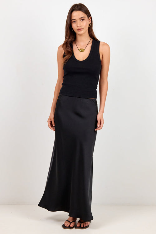 A woman modeling a black sleeveless ribbed tank top and a matching black satin maxi skirt, styled with a gold pendant necklace and brown sandals, against a plain white studio background.