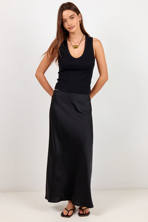 A woman modeling a black sleeveless knit top and a long black satin skirt, paired with black sandals and a gold necklace, against a plain white studio background.