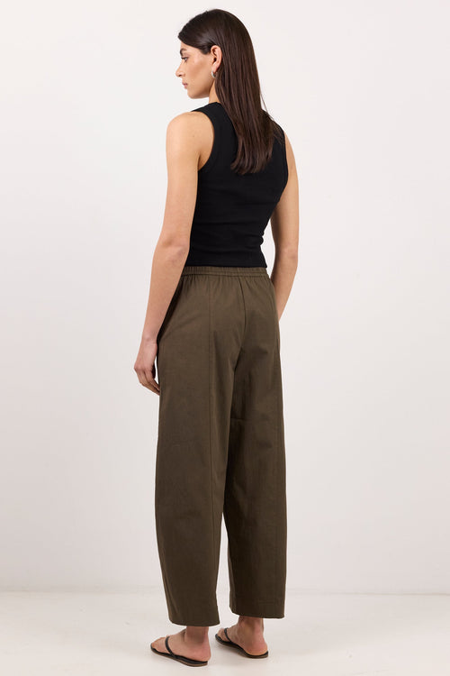 A woman stands in profile wearing wide-leg olive brown trousers, a black sleeveless top, and flat sandals against a plain white studio background.