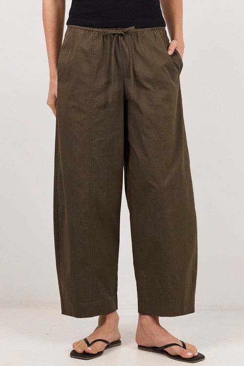 A woman stands against a plain white studio background wearing a black sleeveless top and wide-leg olive green drawstring pants with black sandals.