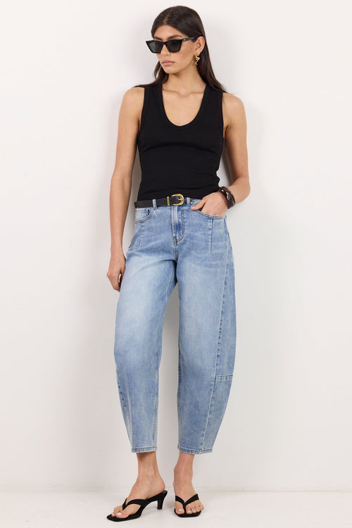 A woman modeling a pair of light-wash, relaxed-fit cropped jeans with a black tank top, black sunglasses, and black sandals against a plain white studio background.