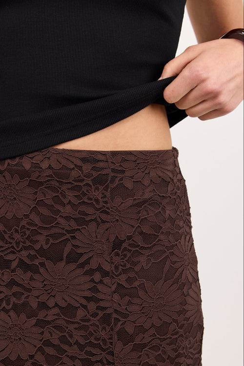 Close-up of a person wearing a dark brown floral lace skirt and lifting the hem of a black ribbed top to show the waist area.