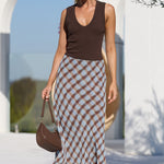 A woman walking outdoors near a white architectural structure and pool, wearing a brown sleeveless top and a long brown-and-beige checkered skirt, with a brown handbag.