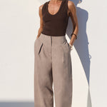 A model stands against a white architectural wall wearing a dark brown ribbed tank top, high-waisted wide-leg taupe trousers, and dark sunglasses.