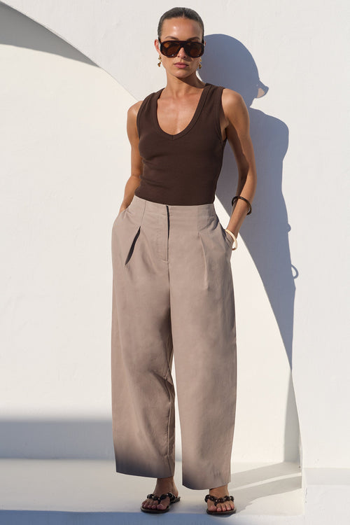 A model stands against a white architectural wall wearing a dark brown ribbed tank top, high-waisted wide-leg taupe trousers, and dark sunglasses.