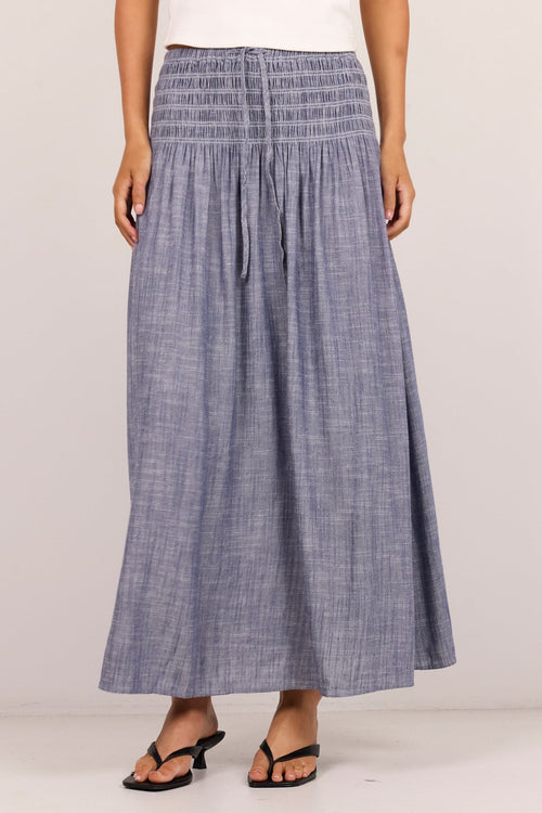 A model wearing a smocked blue-gray linen maxi skirt with a white top and black sandals, standing against a plain off-white studio background.