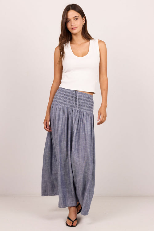A woman modeling a pair of wide-leg, smocked blue-gray pants with a white ribbed tank top and black sandals, standing against a plain light gray studio background.
