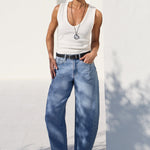 A model wearing a white tank top, wide-leg light blue jeans, a black belt, and black sandals, standing outdoors against a white wall.
