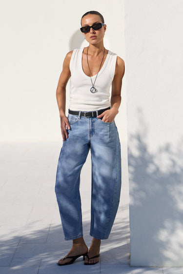 A model wearing a white tank top, wide-leg light blue jeans, a black belt, and black sandals, standing outdoors against a white wall.
