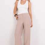 A woman modeling a pair of wide-leg taupe pants with a white tank top and black sandals, standing against a plain white studio background.