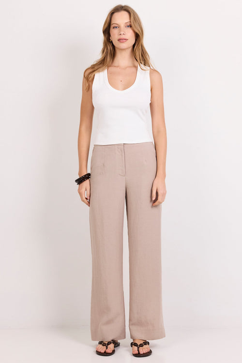 A woman stands against a plain white studio background wearing a white sleeveless top, light beige wide-leg trousers, and dark sandals.