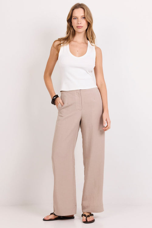 A woman modeling a pair of wide-leg beige trousers with a white tank top and black sandals, standing against a plain white studio background.
