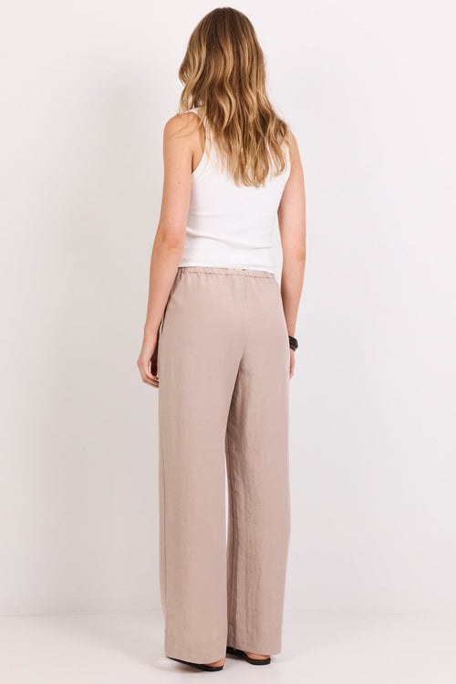 A woman viewed from behind wearing wide-leg beige trousers and a white sleeveless top, standing against a plain white studio background.