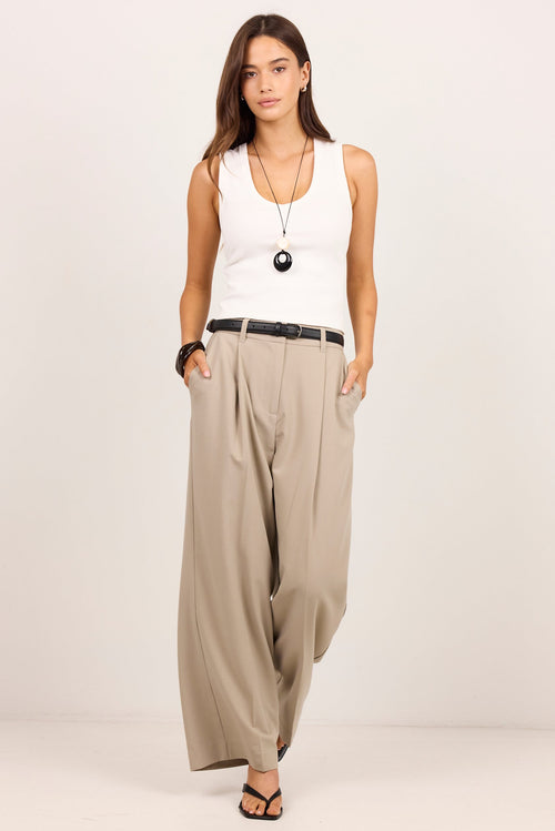 A woman modeling a white sleeveless tank top and beige wide-leg trousers with a black belt, standing against a plain light studio background.