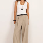 A woman stands against a plain white studio wall wearing a white sleeveless top, wide-leg beige pants, a black belt, and black sandals.