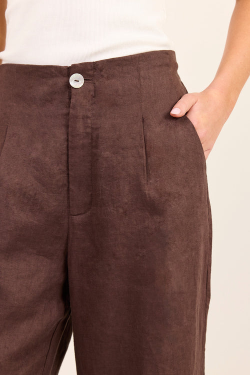 Close-up of a person wearing dark brown wide-leg trousers with a button closure and side pockets, paired with a white top against a light beige background.