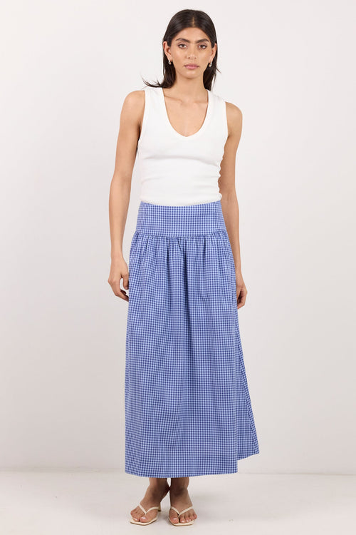 A woman modeling a white sleeveless tank top and a high-waisted, blue-and-white gingham pleated maxi skirt, standing against a plain off-white studio background.