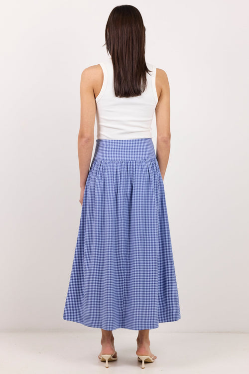 A woman viewed from behind wearing a sleeveless white top and a blue-and-white gingham pleated midi skirt, standing against a plain light gray studio background.