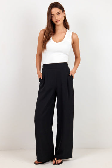 A woman modeling a white sleeveless tank top and black wide-leg trousers against a plain white studio background.