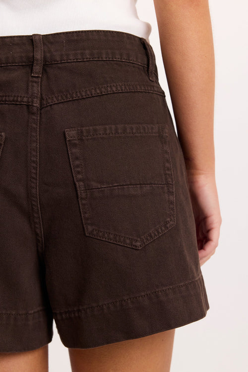 Close-up back view of a person wearing dark brown denim shorts with back patch pockets, shown against a plain light background.