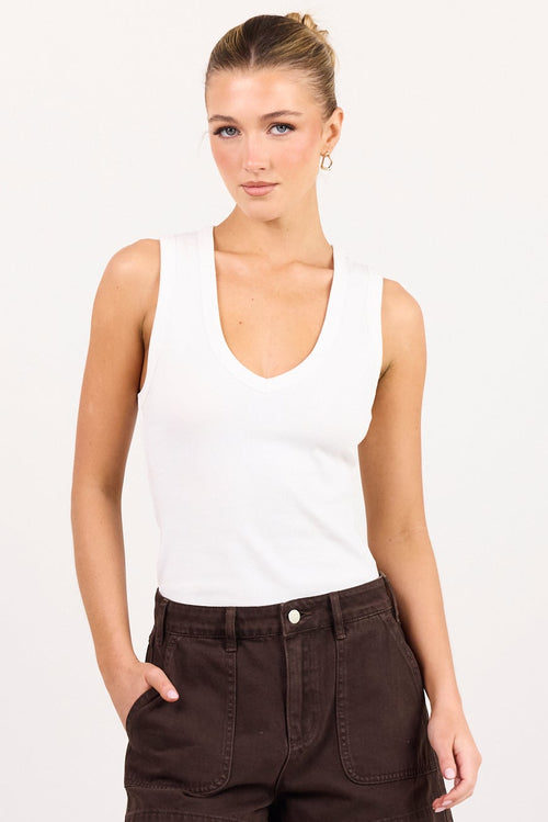 A woman modeling a white sleeveless tank top and dark brown shorts against a plain white studio background.