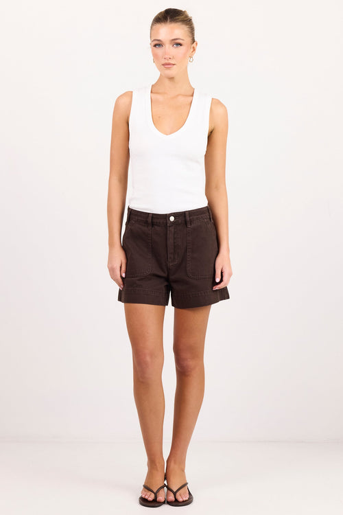 A woman modeling a white ribbed tank top and dark brown shorts against a plain white studio background.