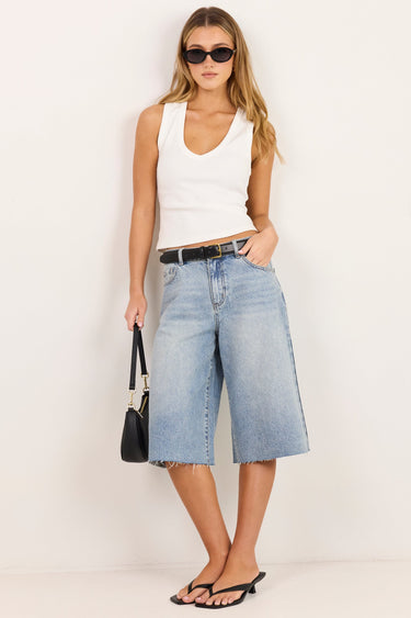 A woman modeling a pair of light-wash blue denim culotte jeans with a black belt, paired with a white tank top, black sunglasses, and black sandals, standing against a plain white studio background.