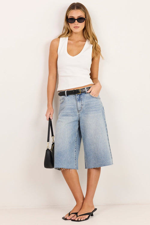 A woman modeling a pair of light-wash blue denim culotte jeans with a black belt, paired with a white tank top, black sunglasses, and black sandals, standing against a plain white studio background.