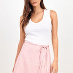 A woman modeling a light pink wrap mini skirt with a tie waist, paired with a white tank top, against a plain white studio background.