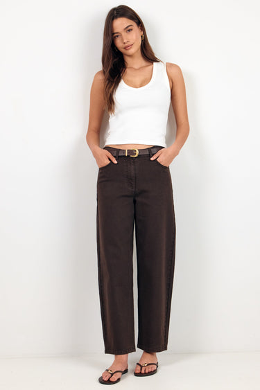 A woman modeling dark brown wide-leg cropped pants with a black belt, paired with a white tank top and black sandals, standing against a plain white studio background.