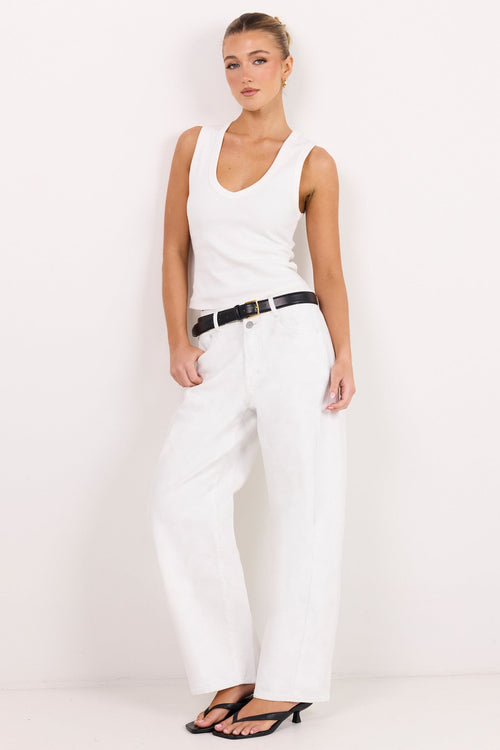A woman modeling a white sleeveless tank top and white wide-leg jeans with a black belt, standing against a plain white studio background.