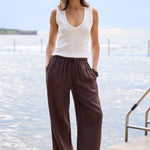 A woman stands on a stone pier by the water wearing a white sleeveless V-neck top and dark brown wide-leg pants, with a metal ladder visible to her right.