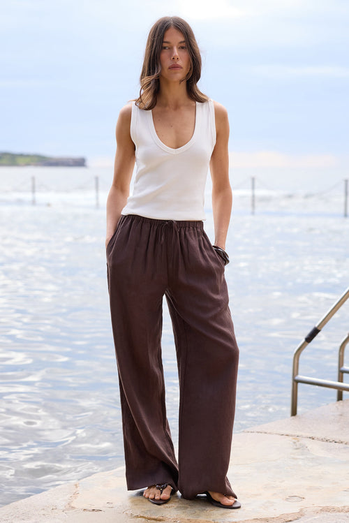 A woman stands on a stone pier by the water wearing a white sleeveless V-neck top and dark brown wide-leg pants, with a metal ladder visible to her right.