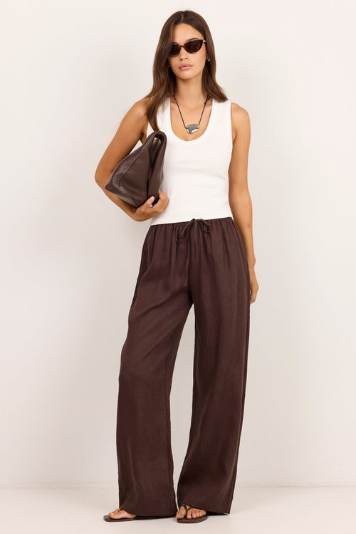 A woman modeling wide-leg brown pants with a white tank top, sunglasses, and a dark brown handbag against a plain white studio background.